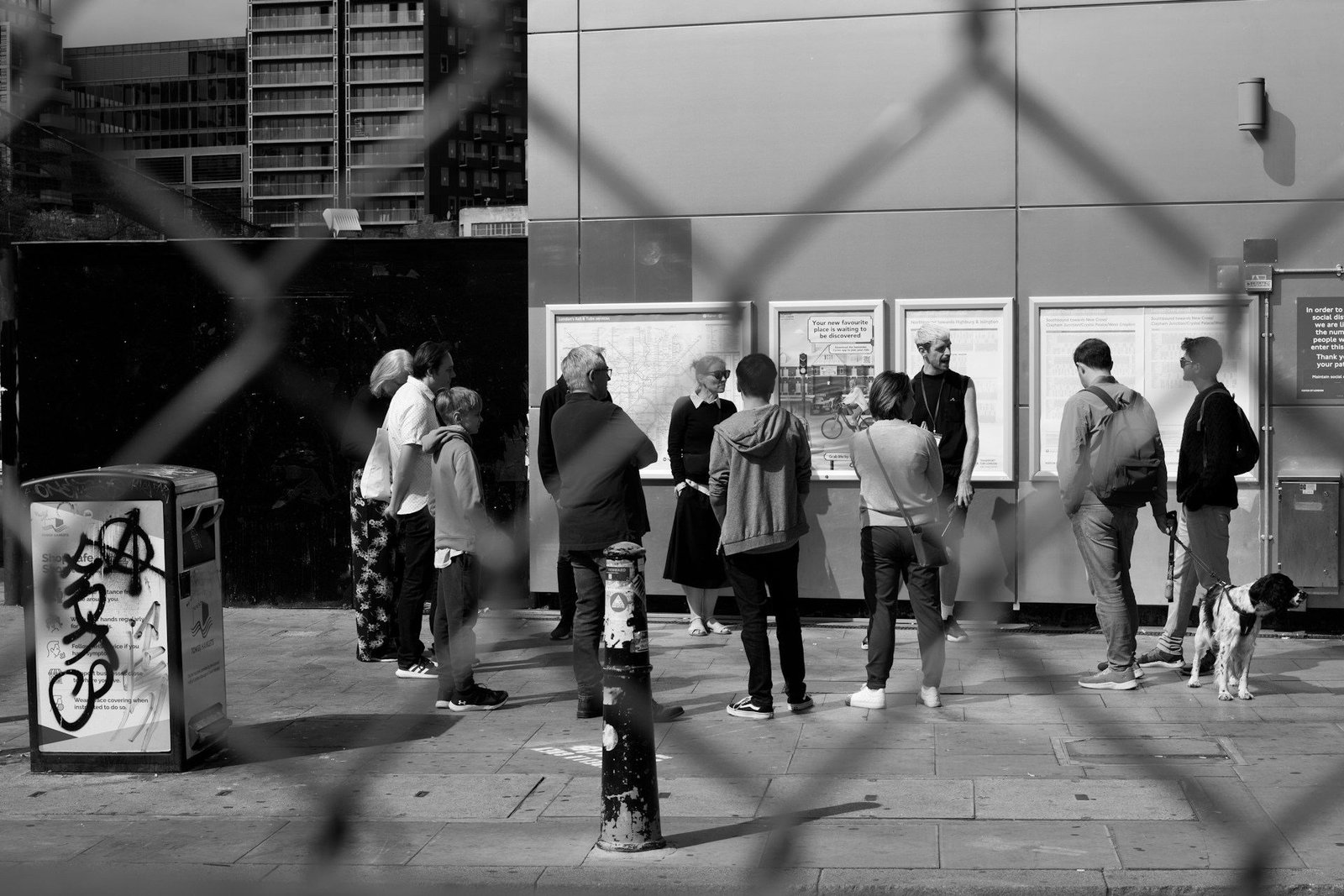 People reading a street noticeboard, seen through a wire fence